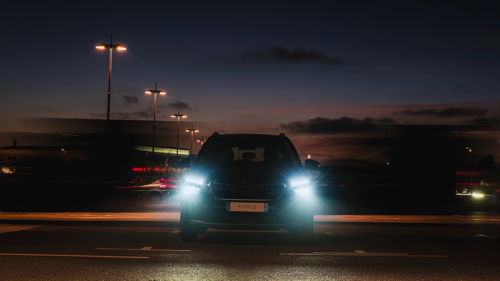 Skoda Kamiq de face, phares allumés, au crépuscule parmi les lumières de la ville.