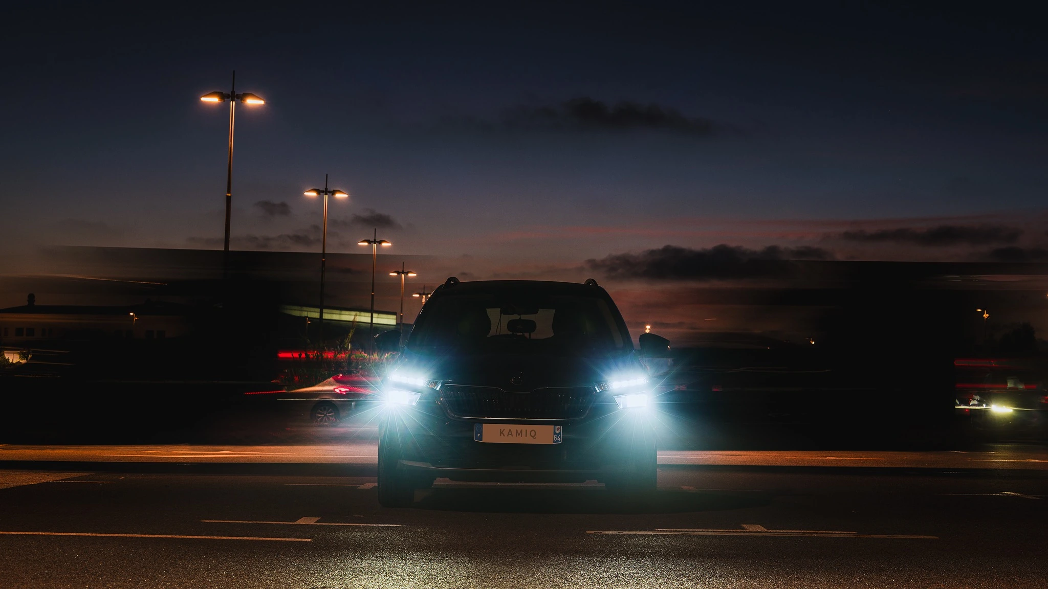 Skoda Kamiq de face, phares allumés, au crépuscule parmi les lumières de la ville.